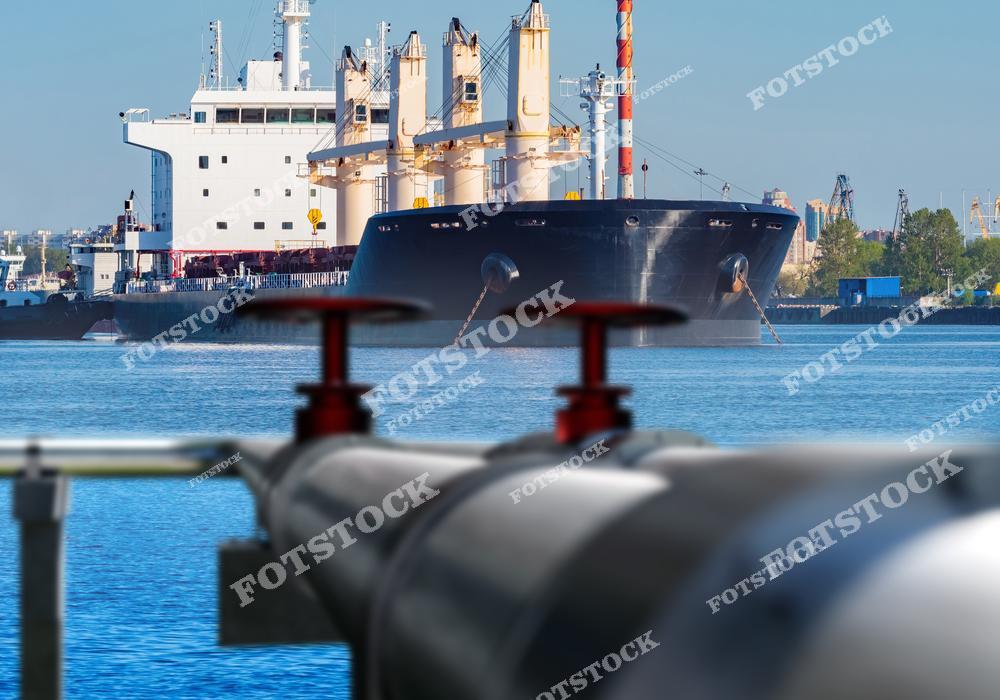 На изображении крупный нефтяной танкер, пришвартованный в порту, с трубопроводами и вентилями на переднем плане. Фотография подчёркивает важность морской инфраструктуры для транспортировки нефти и сырьевых ресурсов. Это иллюстрация взаимодействия логистики и энергетической промышленности.